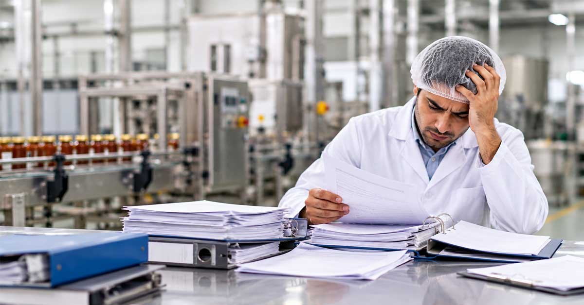 SMETA 7.0 Management Systems Assessment (MSA): What Auditors Now Check 1 Stressed factory manager in white coat reviewing piles of audit documents at desk with production line in background